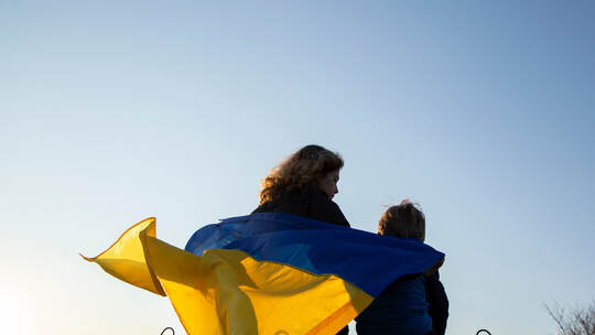little boy and a woman sit with their backs at sunset against sky with a blue and yellow Ukrainian flag